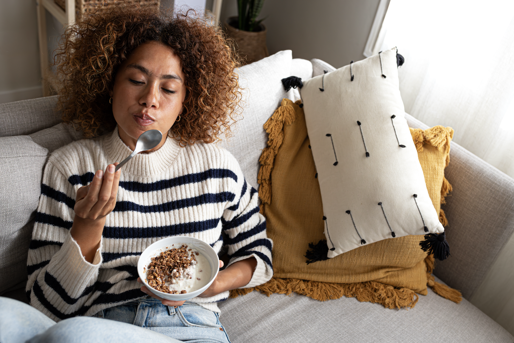 A woman is seated on a couch, wearing a striped sweater and eating a bowl of granola with yogurt. There are pillows beside her