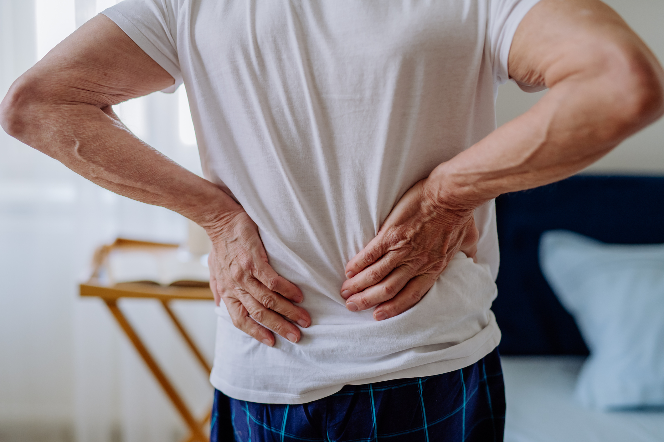 Person holding their lower back, suggesting back pain, while standing in a bedroom wearing a white t-shirt and checkered pants
