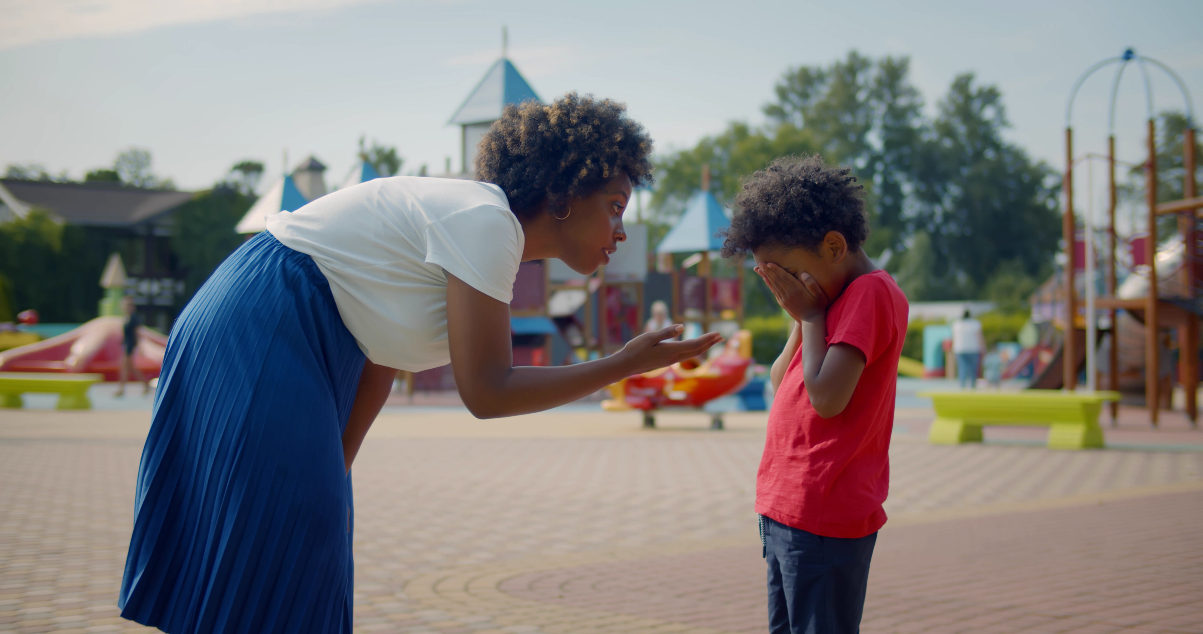 A woman in a white top and blue skirt bends down, talking to a child who is covering their face in a playground
