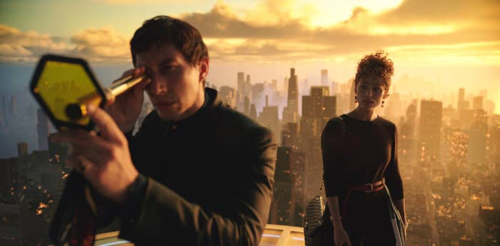 Adam Driver using a telescope, while Nathalie Emmanuel stands behind him. Skyscrapers and a golden sky form the background