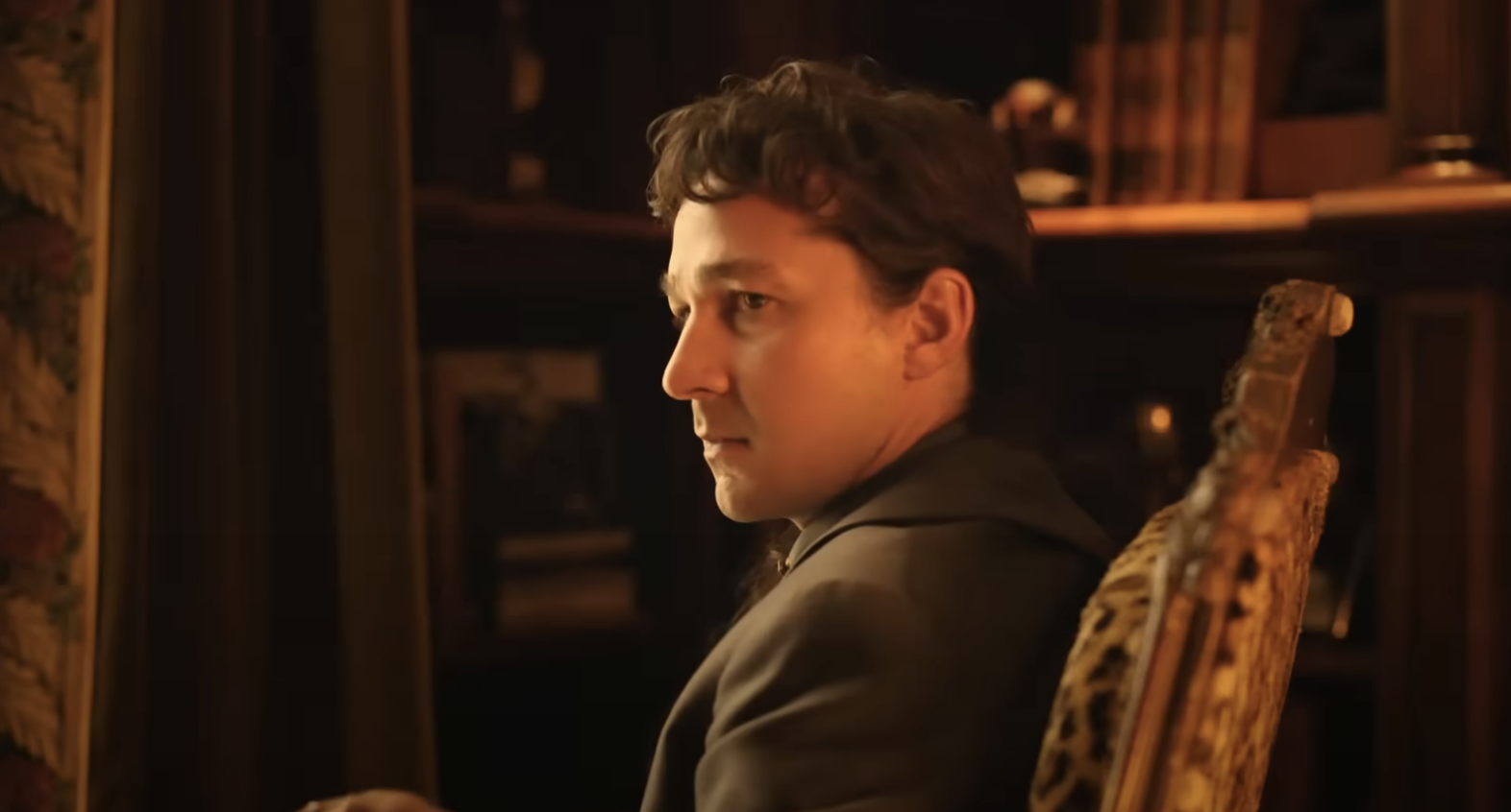 Shia is sitting in an ornate chair, wearing a formal suit, with a contemplative expression, and bookshelves in the background in a scene from the film