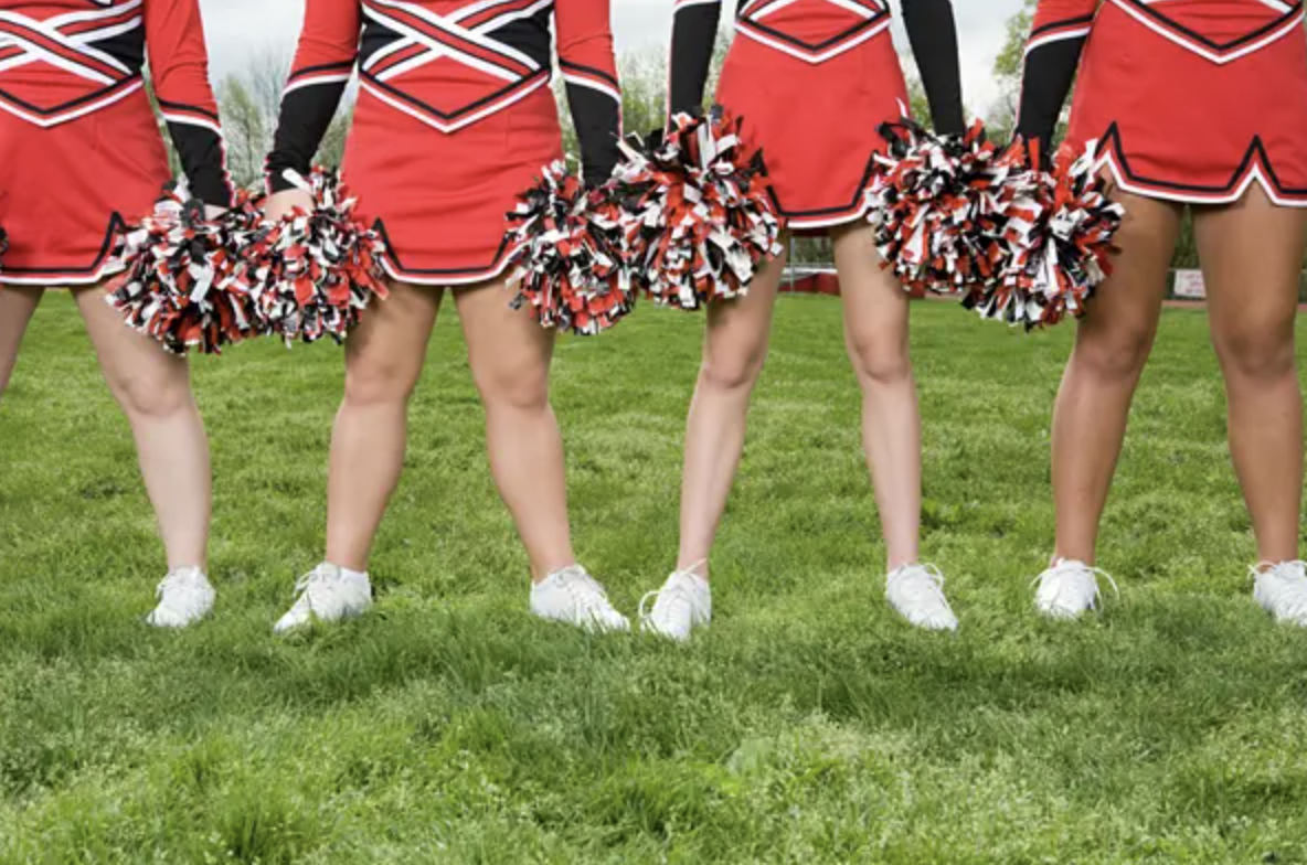Four cheerleaders in uniform stand on grassy field holding pom-poms. Faces are not visible, focusing on legs, uniforms, and pom-poms