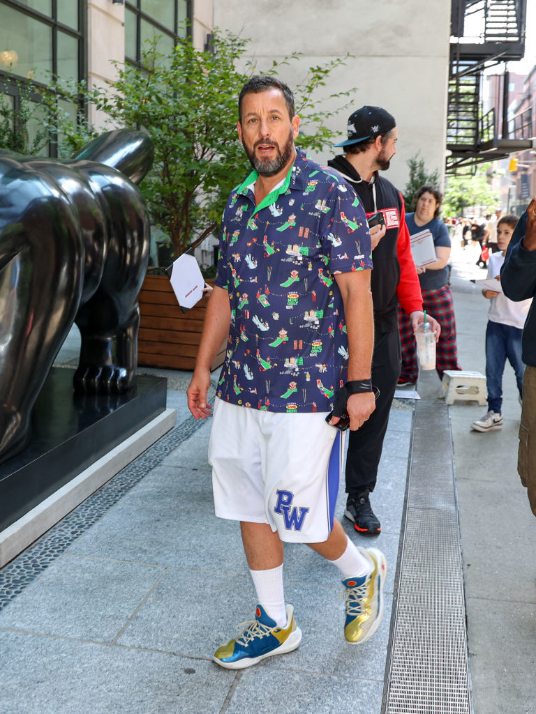 Adam Sandler walking on a city sidewalk wearing a patterned shirt, basketball shorts, and sneakers. A man in a tracksuit follows behind him
