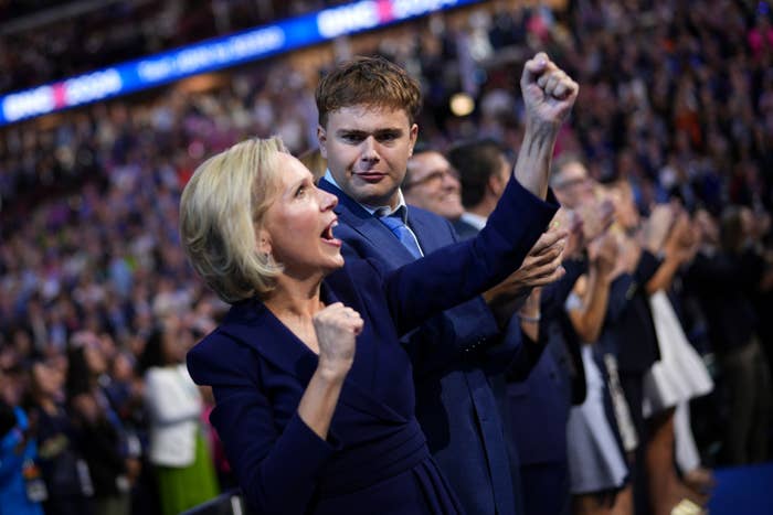 Kimberly Guilfoyle energetically raises her fist in celebration, wearing a formal dress. Eric Trump stands behind, also dressed formally, in a crowded venue