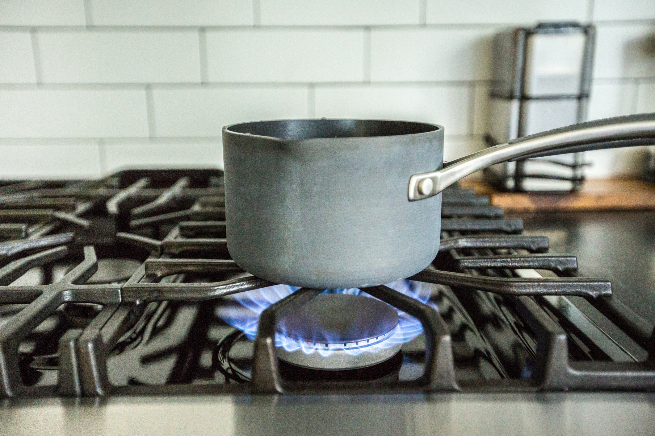 A metal saucepan heats on a gas stove with visible blue flames. The kitchen background includes a tiled wall and a counter with objects