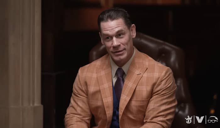 John Cena in a plaid suit, seated, speaking during an interview