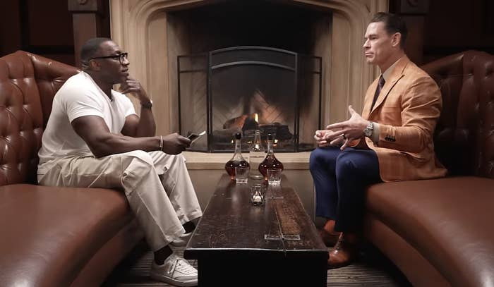 Shannon Sharpe and John Cena sit on leather couches for an interview by a fireplace, with bottles and glasses on the table between them