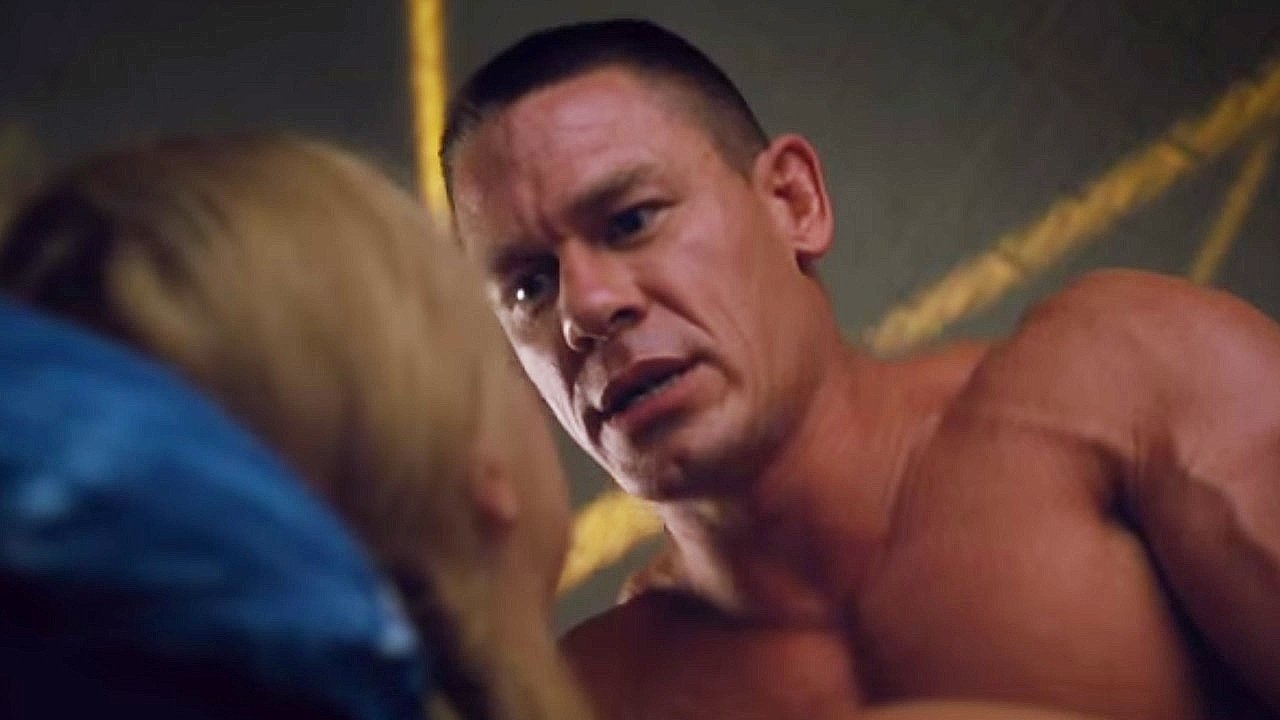 John Cena, shirtless, leans forward with a serious expression while looking at a blonde woman whose face is out of frame