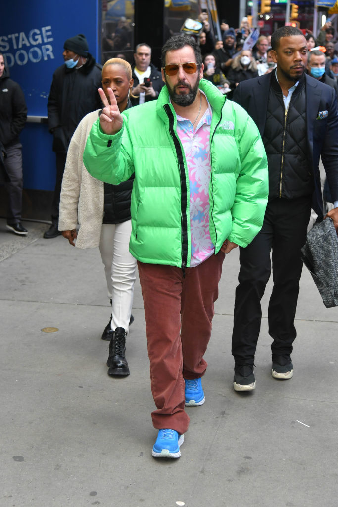 Adam Sandler on a city street, wearing a bright puffer jacket, patterned shirt, brown pants, and blue sneakers, making a peace sign gesture