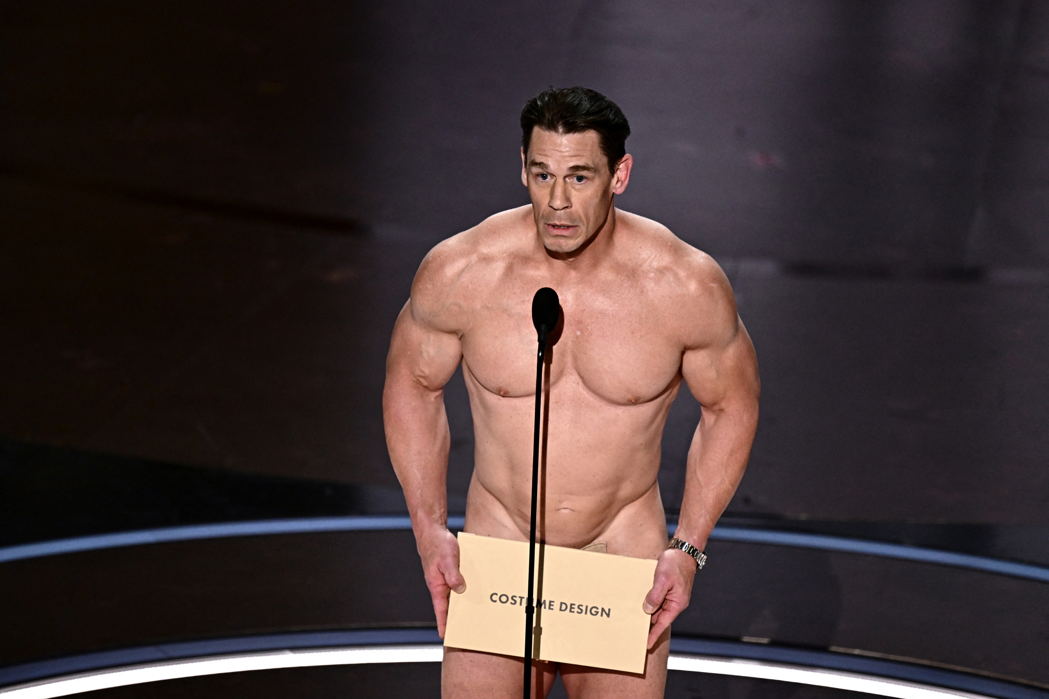 John Cena on stage, humorously dressed in minimal attire, holding an award envelope at an event