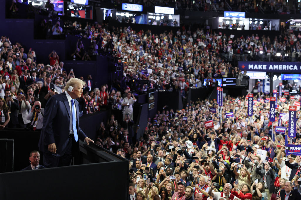 Donald Trump stands on a platform at an event, surrounded by a crowd of supporters holding signs and banners