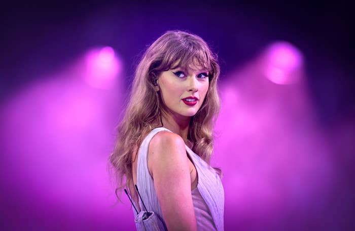 Taylor Swift stands on stage, looking over her shoulder, wearing a sleeveless dress, with a backdrop of purple stage lights