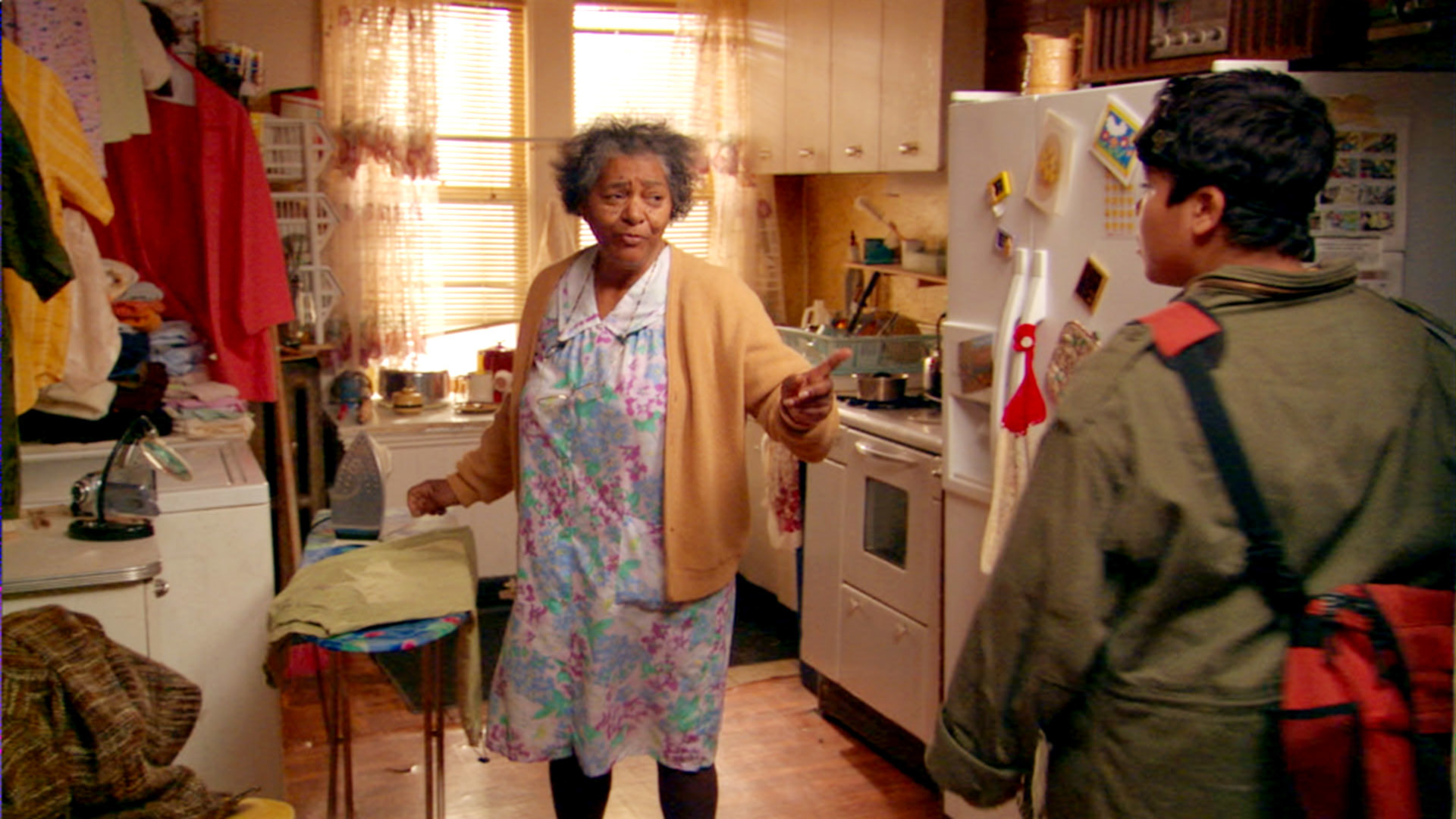 An older woman in a floral dress and cardigan talks to a young man with a backpack in a cluttered kitchen