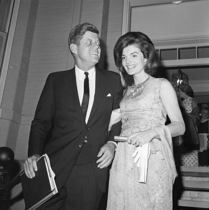 John F. Kennedy in a suit and tie, and Jacqueline Kennedy in a sleeveless, elegant dress holding gloves, smiling and walking together