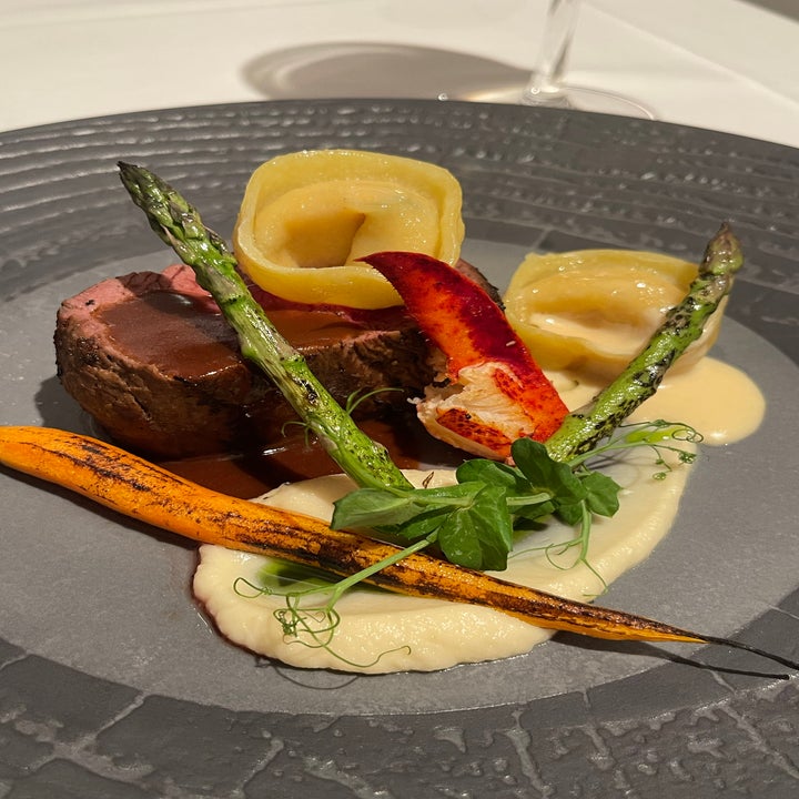 A gourmet dish featuring beef tenderloin, lobster claw, asparagus, baby carrot, ravioli, and a potato puree, beautifully plated on a dark plate