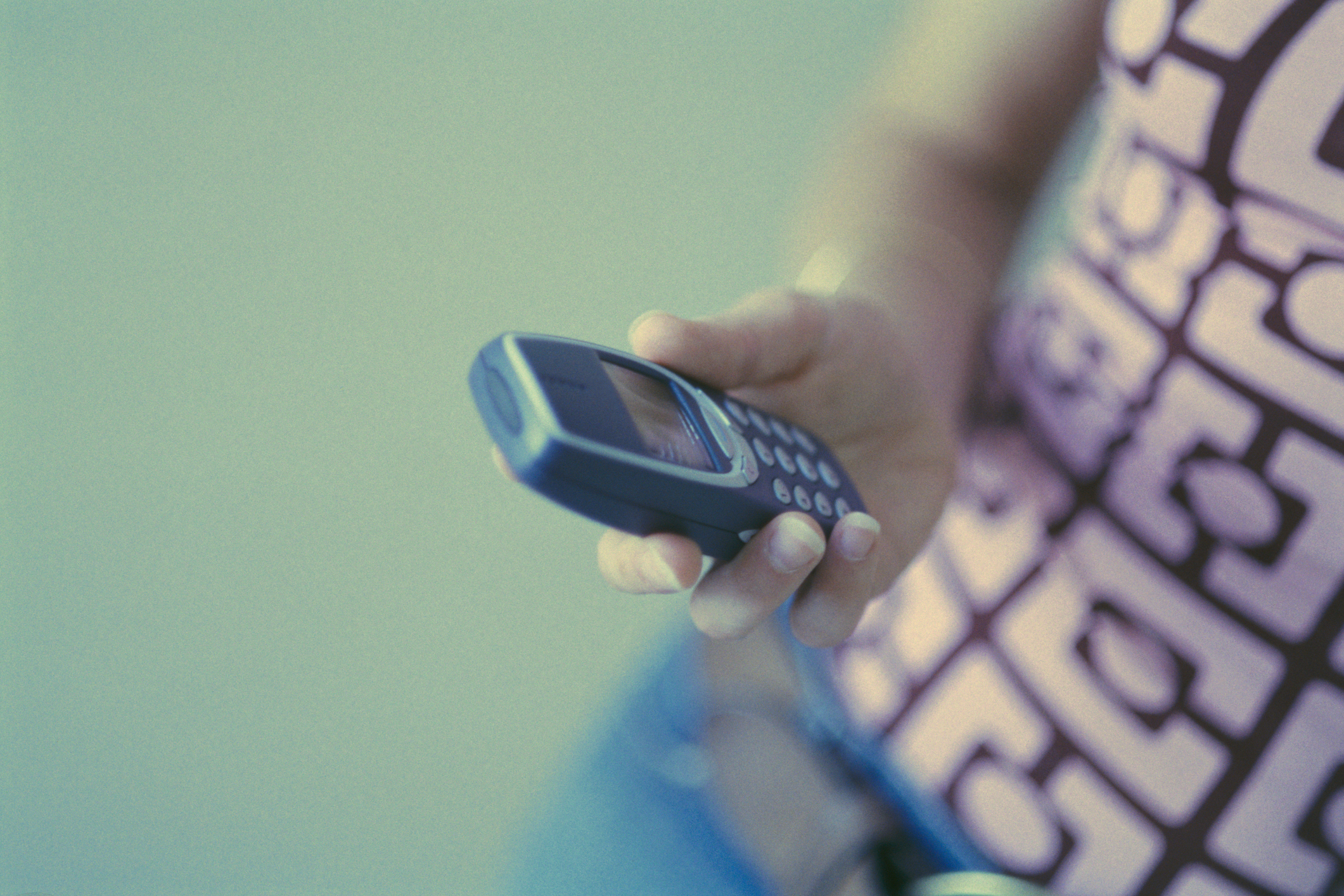 A hand holds a vintage mobile phone. The person is wearing a patterned top