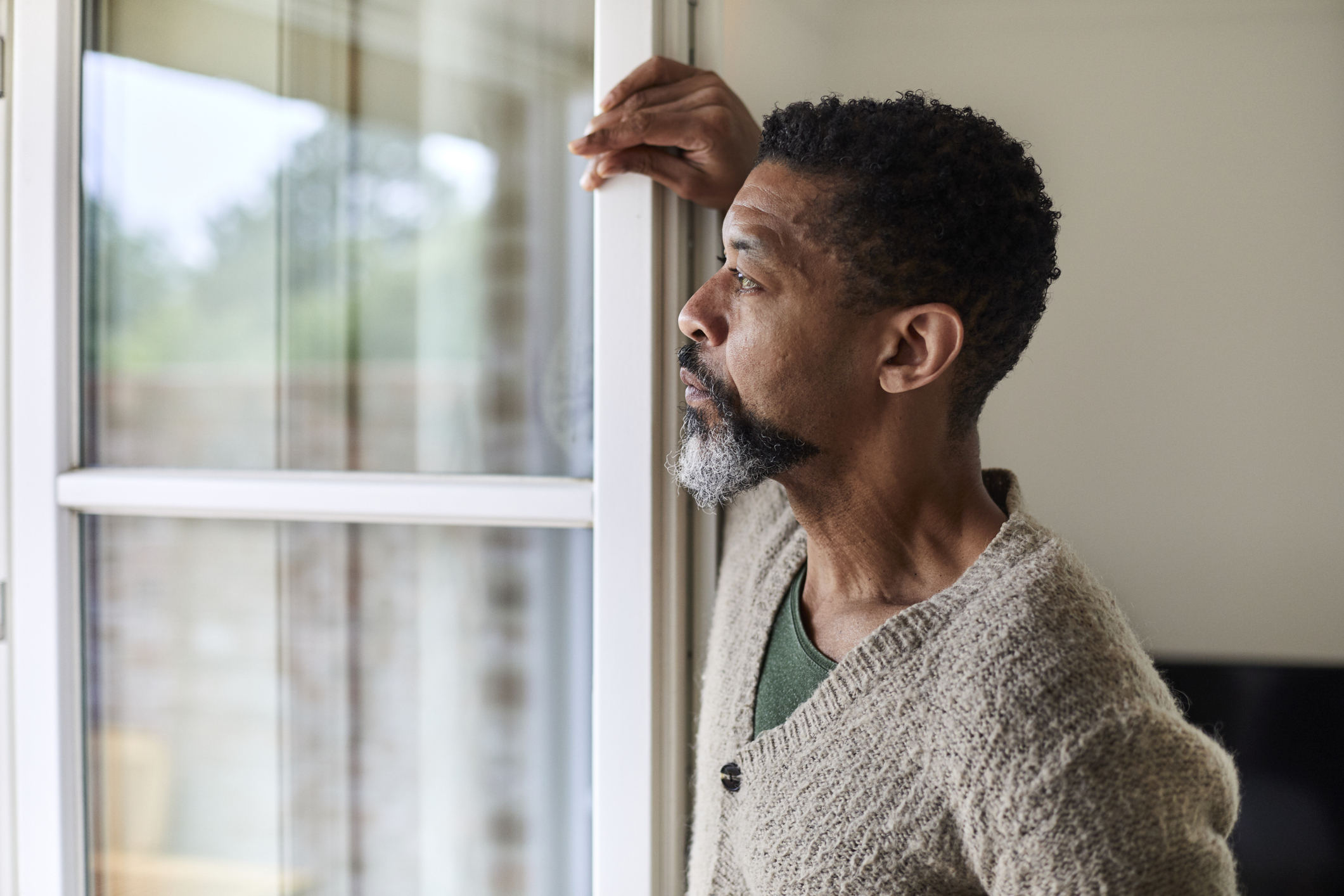 A contemplative man wearing a knitted sweater leans against a window, gazing outside