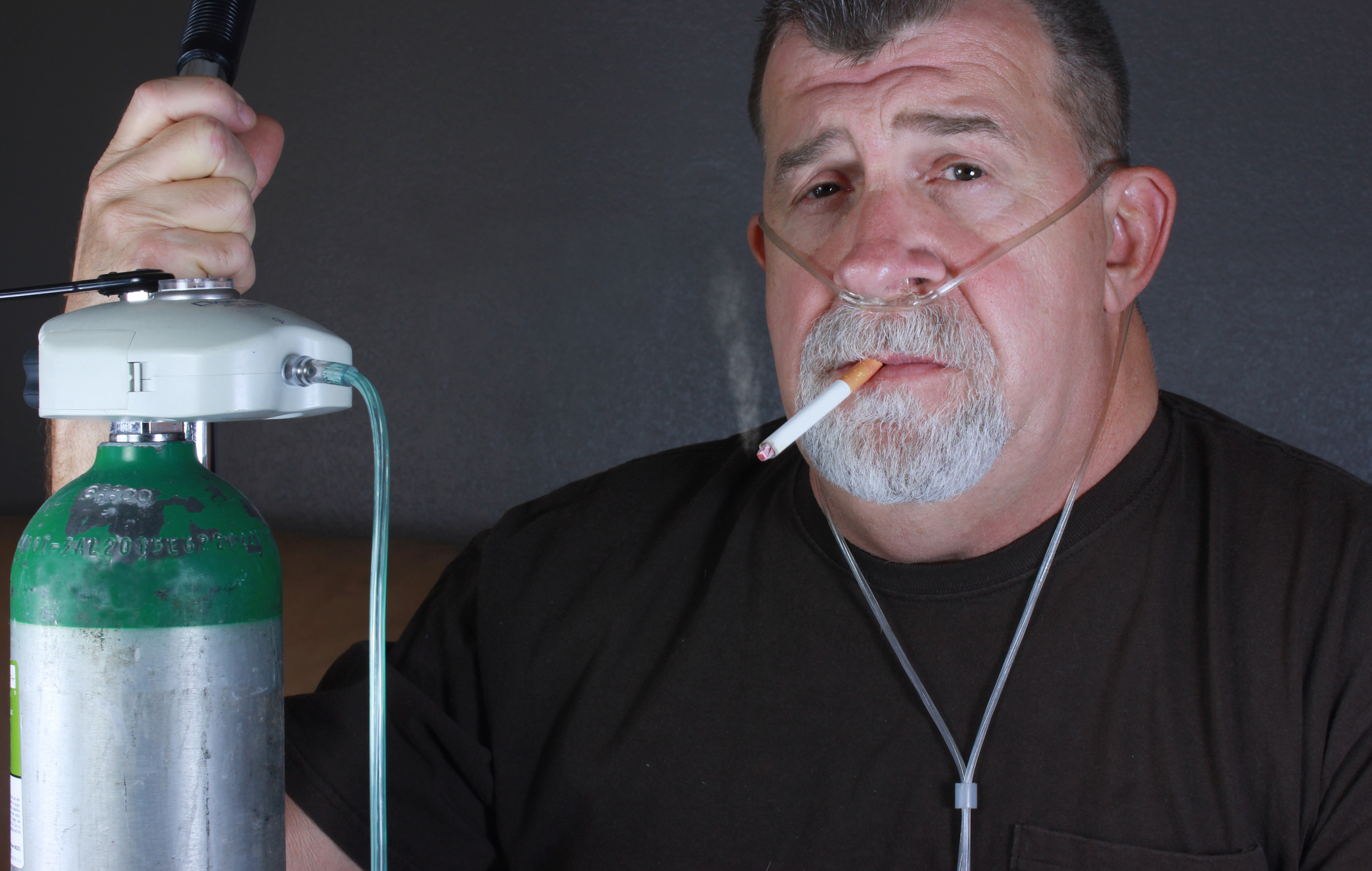 A man with a nasal oxygen tube smokes a cigarette while holding an oxygen tank