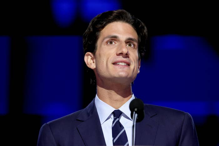 Jack in a suit and tie is standing at a microphone, smiling slightly, with a blurred background behind him. Names are not included for privacy