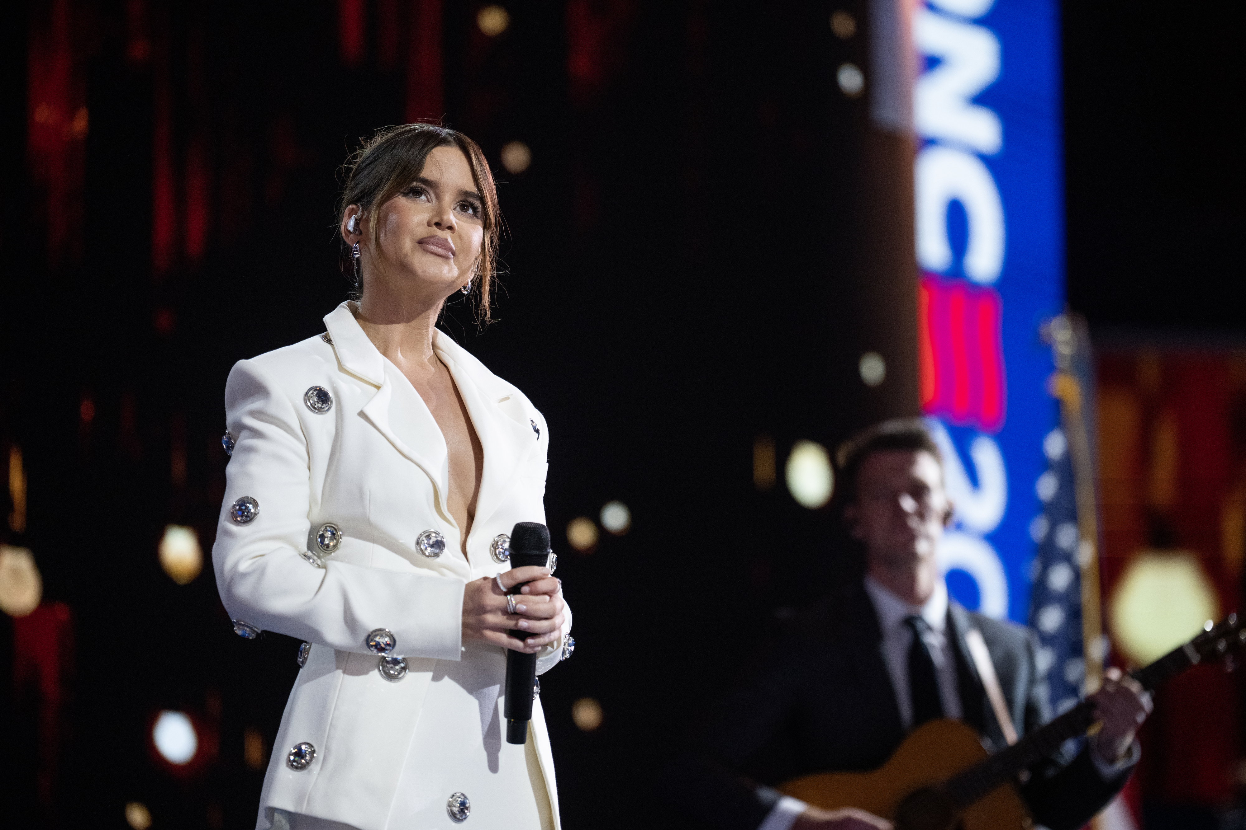 Maren Morris performing on stage in a stylish white outfit with jeweled embellishments, holding a microphone; a guitarist plays in the background