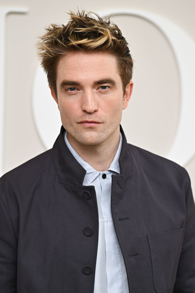 Robert Pattinson wearing a black jacket over a light shirt, posing in front of a light backdrop with large text partially visible behind him