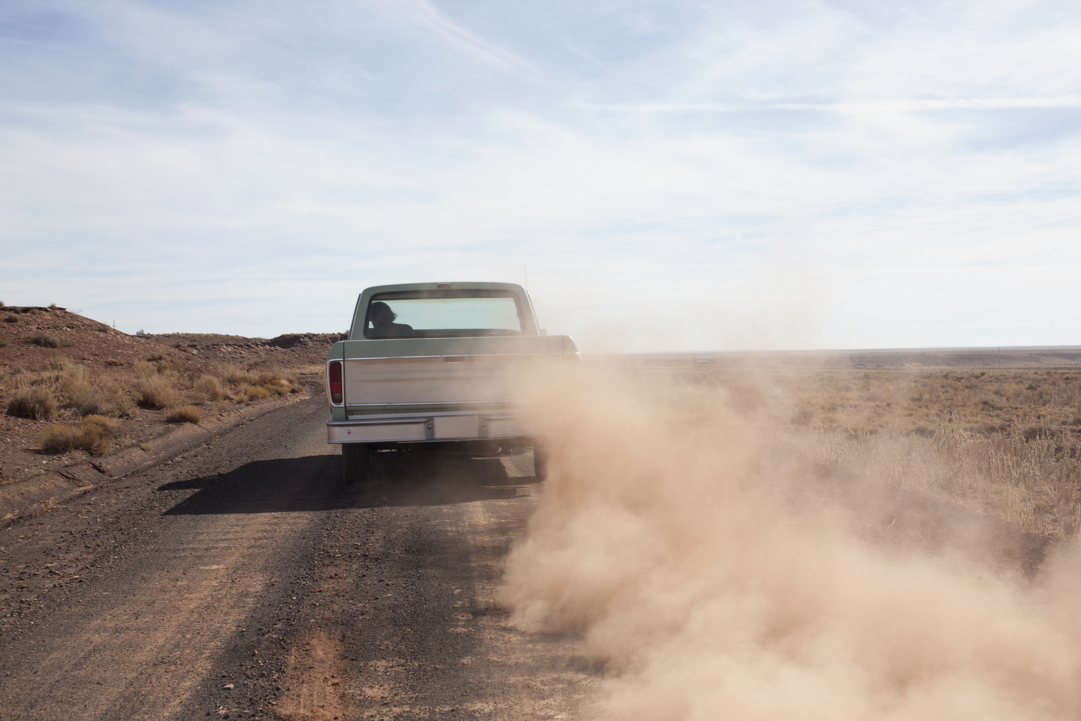 A pickup truck drives down a dusty desert road emitting a cloud of dust behind it. The scene is barren with clear skies