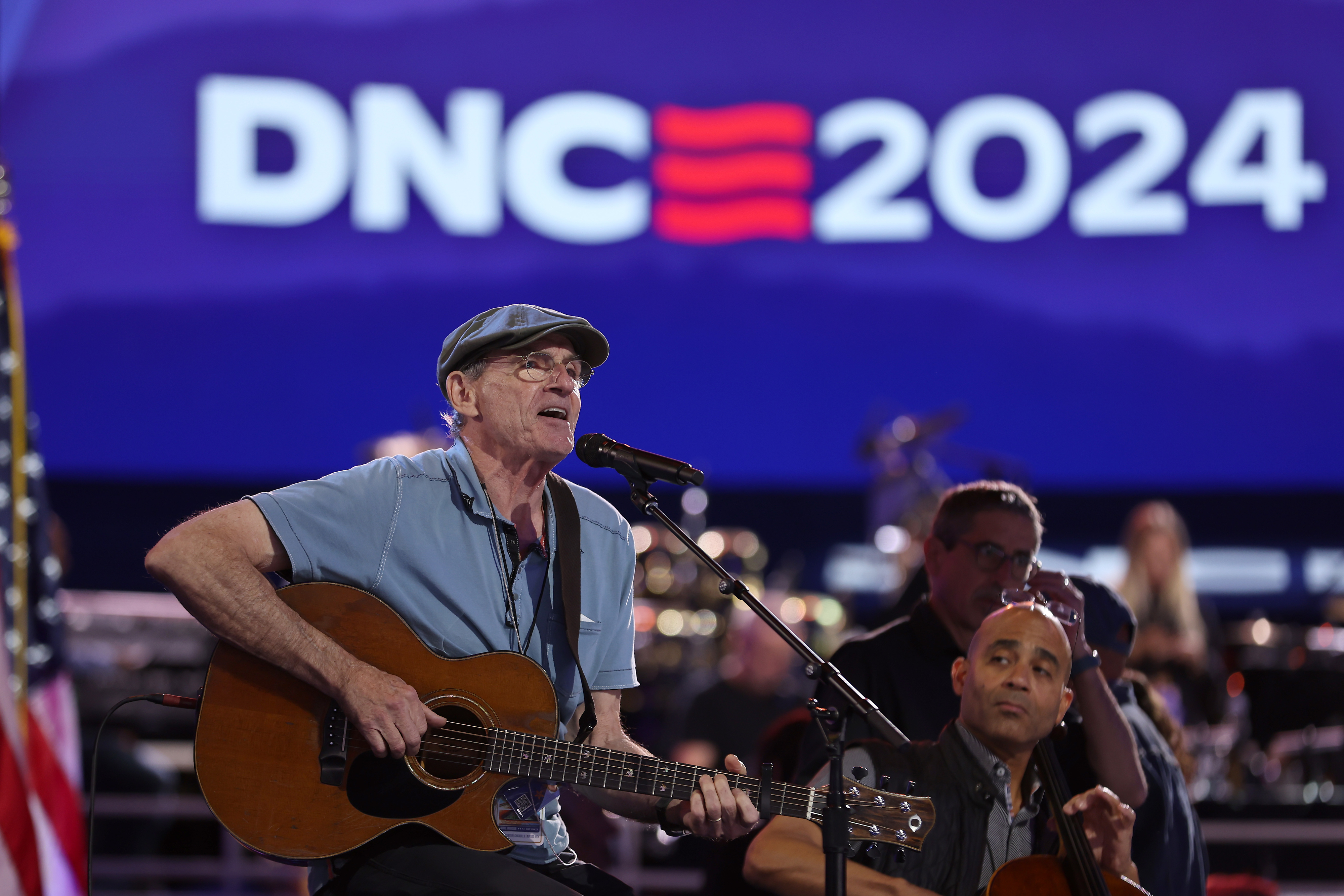 James Taylor performs with an acoustic guitar on stage at the DNC 2024 event. Two additional musicians play in the background