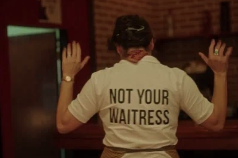 A person stands with their back to the camera, wearing a shirt that reads &quot;NOT YOUR WAITRESS.&quot; Their hands are raised