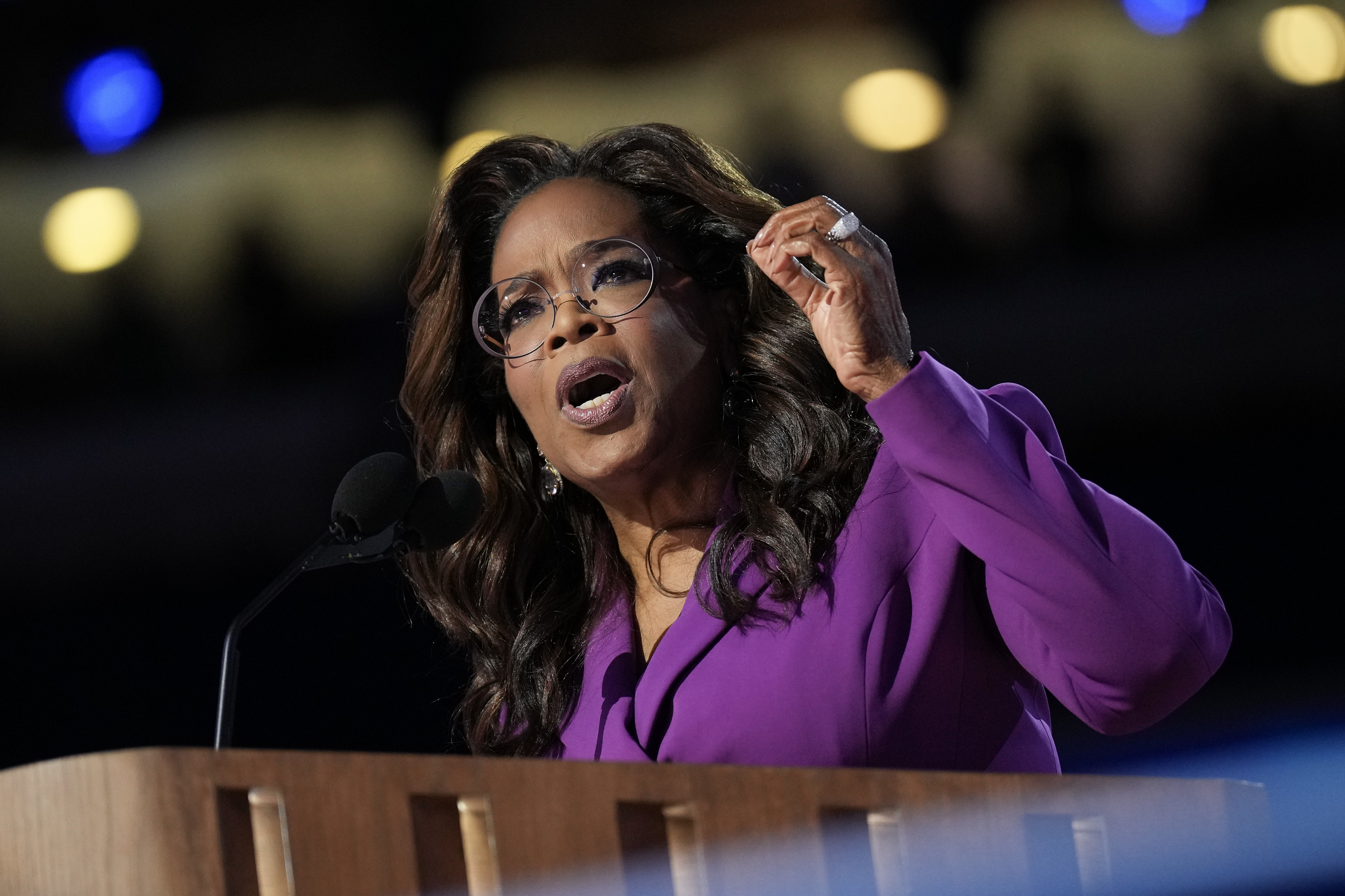 Oprah Winfrey stands at a podium speaking passionately while gesturing with her hand. She wears glasses and a formal outfit