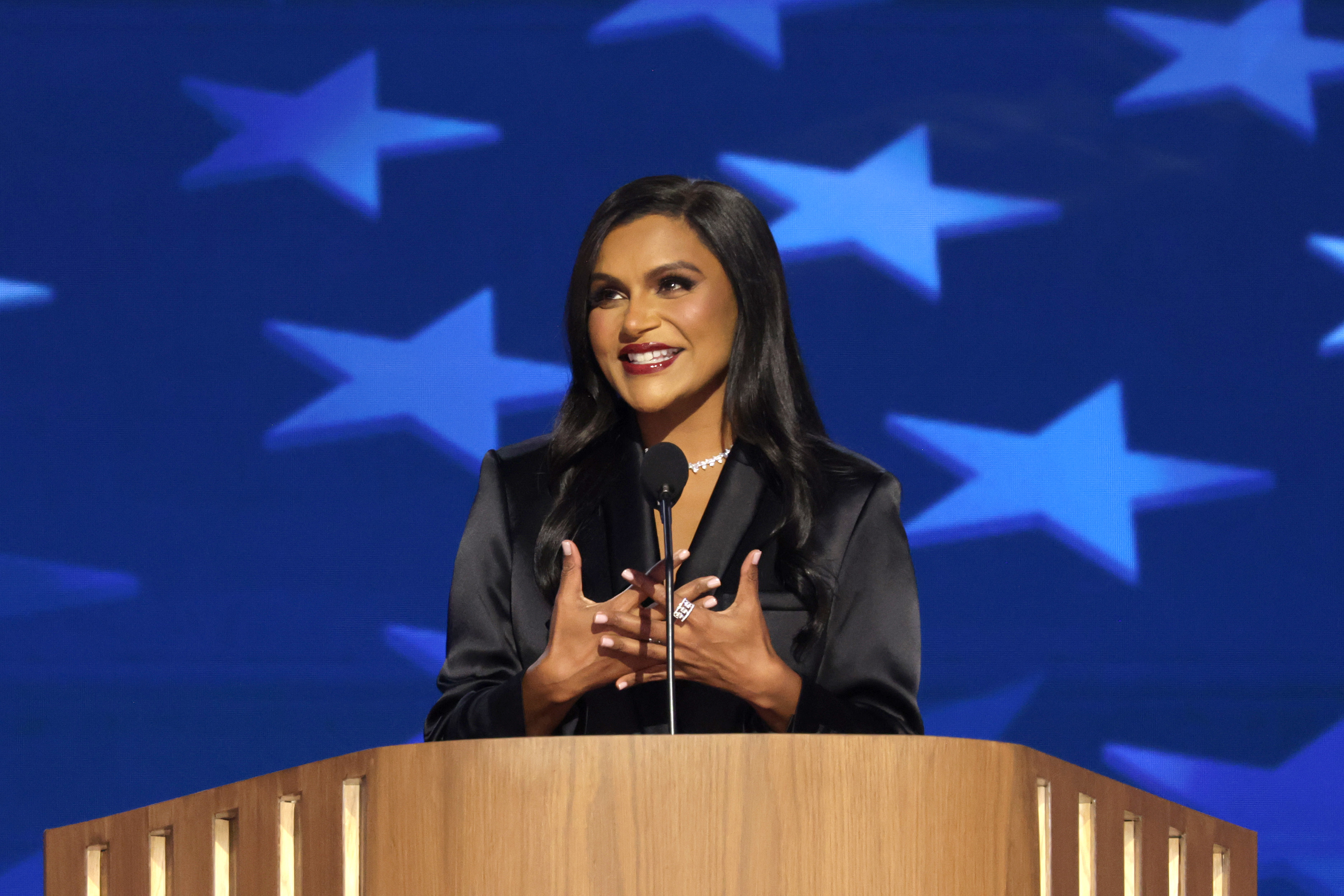 Mindy Kaling speaks at a podium, gesturing with her hands, against a backdrop of star motifs. She is wearing a sleek, elegant outfit