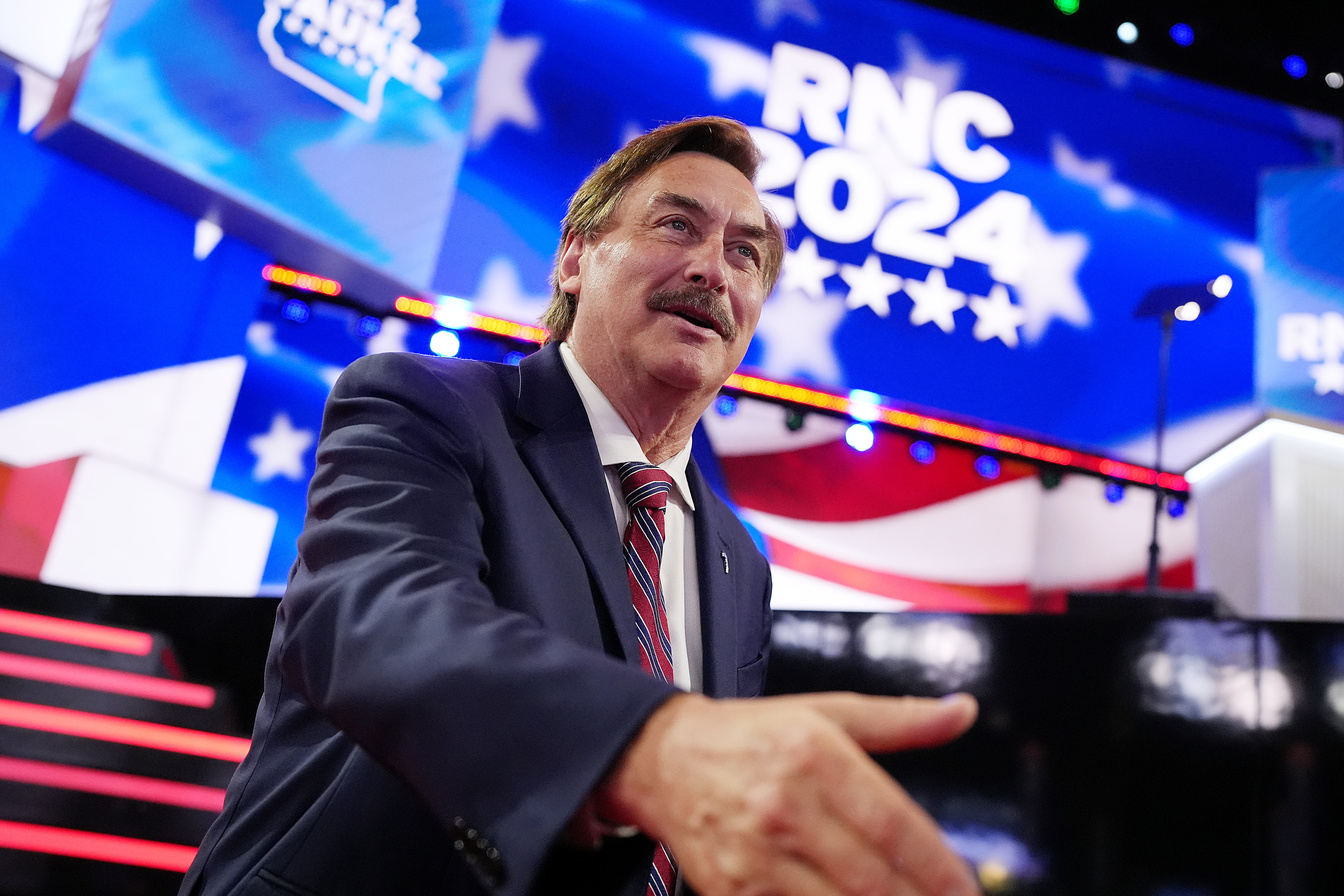 Mike Lindell extends his hand at the RNC 2024 event stage. He is wearing a suit and tie. The background features American flag elements and &quot;RNC 2024&quot; text