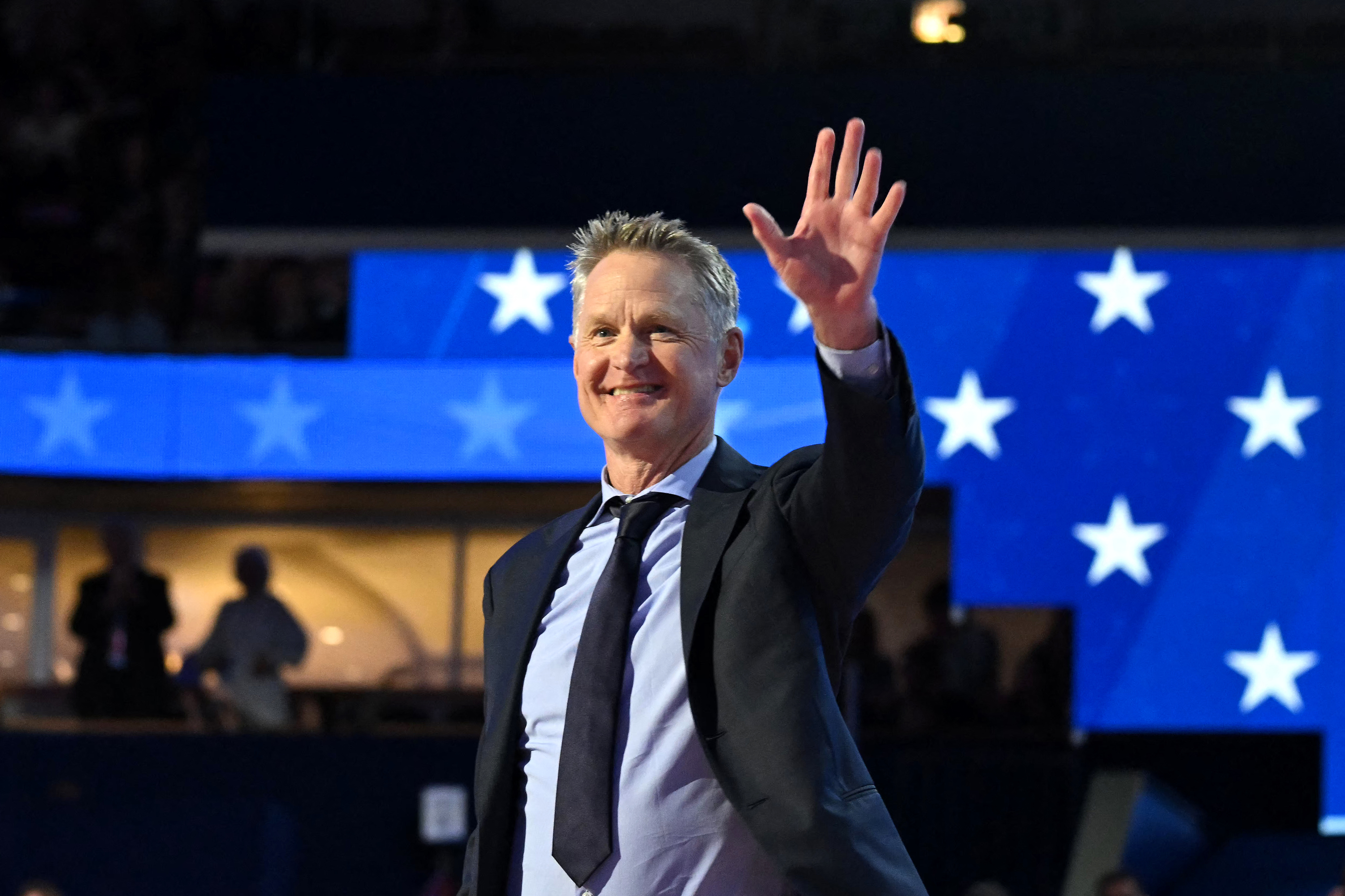 Steve Kerr waves while wearing a suit and tie in front of a backdrop with stars