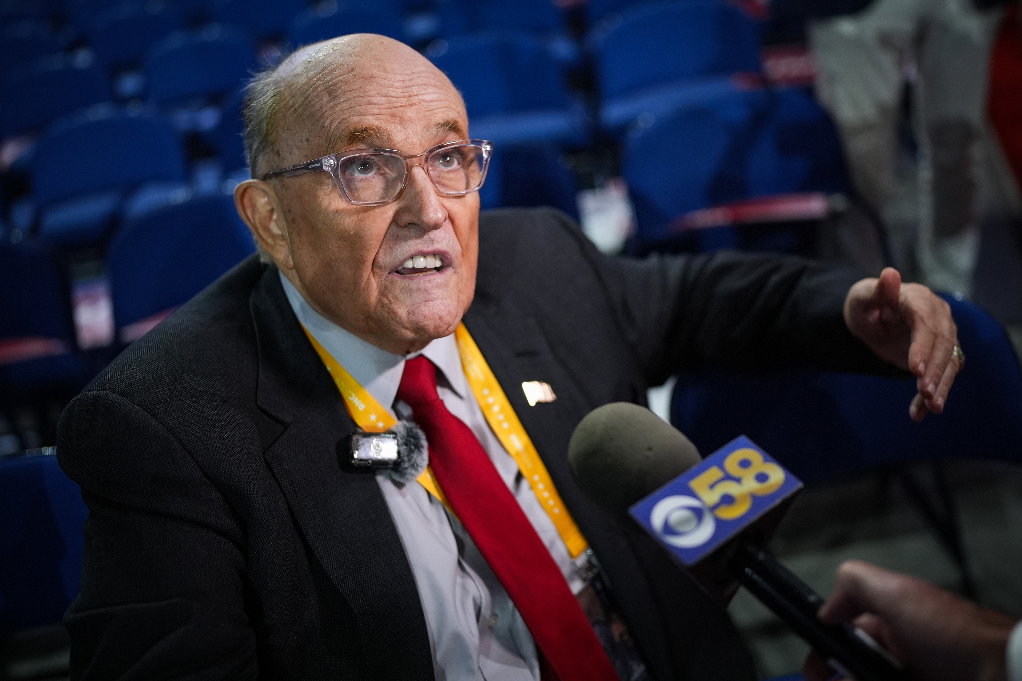 Rudy Giuliani speaks into several microphones at an event, wearing a suit with a red tie and a lanyard badge