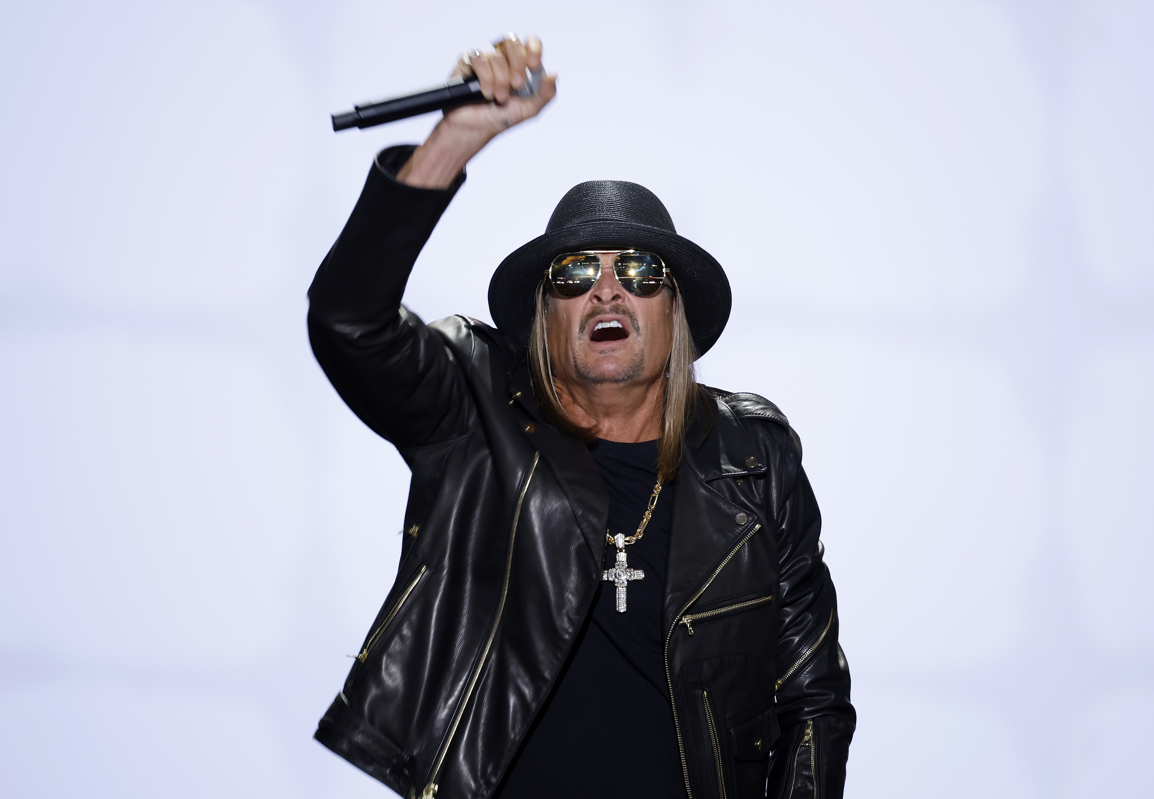 Kid Rock performs on stage wearing a black leather jacket, black hat, and sunglasses, holding a microphone
