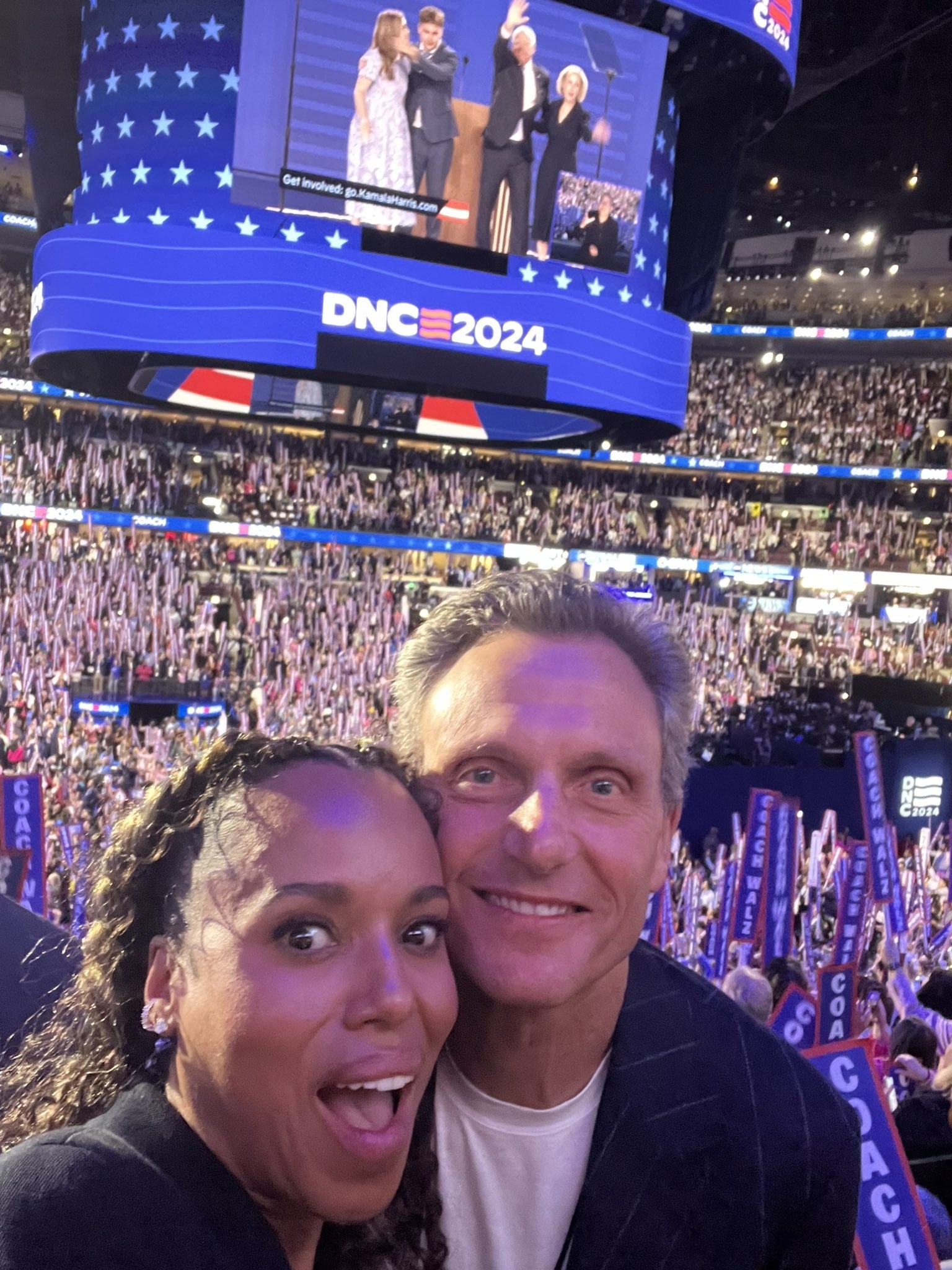 Kerry Washington and Tony Goldwyn take a selfie at the DNC 2024 convention. A large crowd and a big screen showing presenters on stage are visible in the background