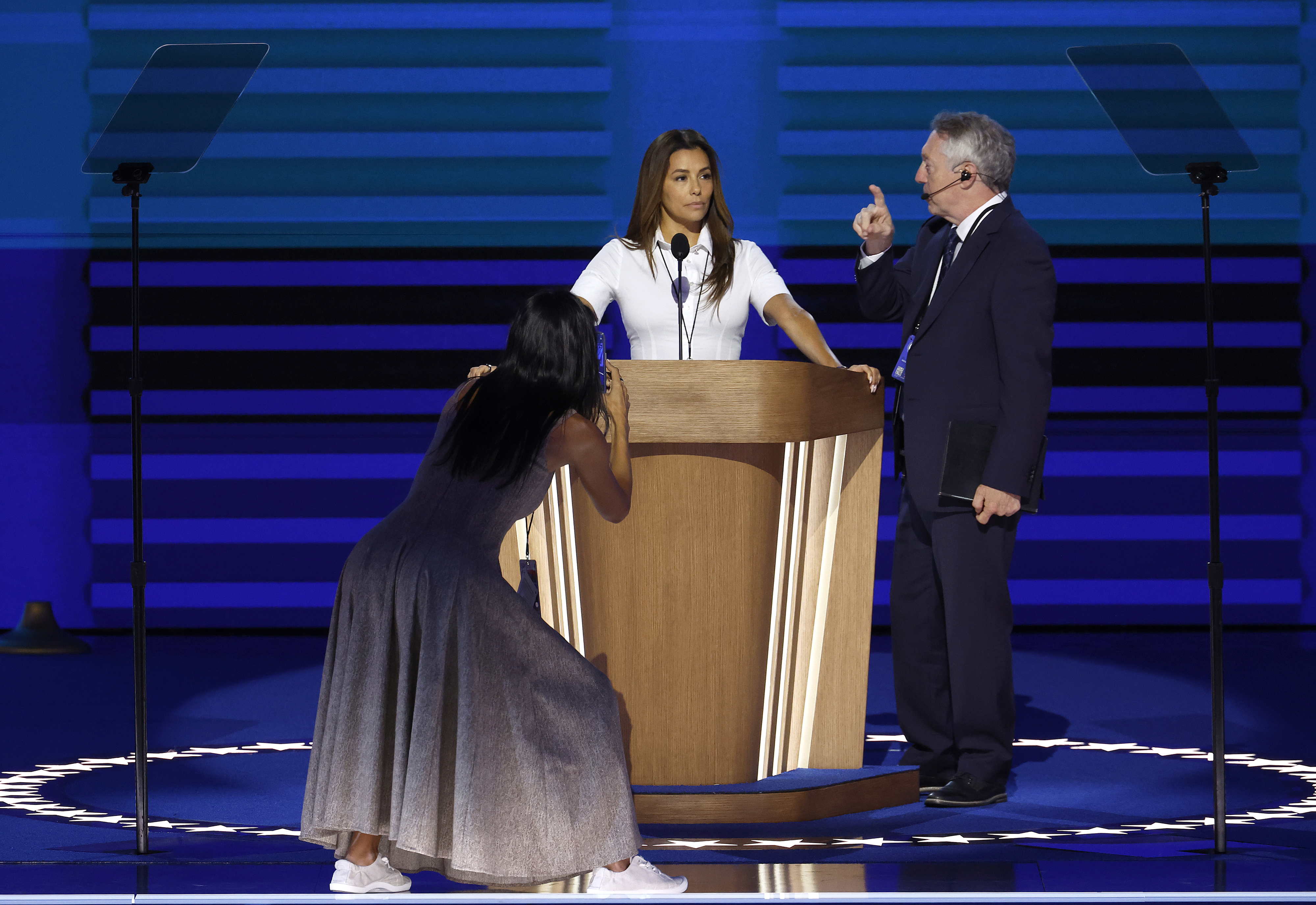 Mindy Kaling stands behind a podium speaking, with Regina King kneeling and taking a photo, and a man in a suit gesturing