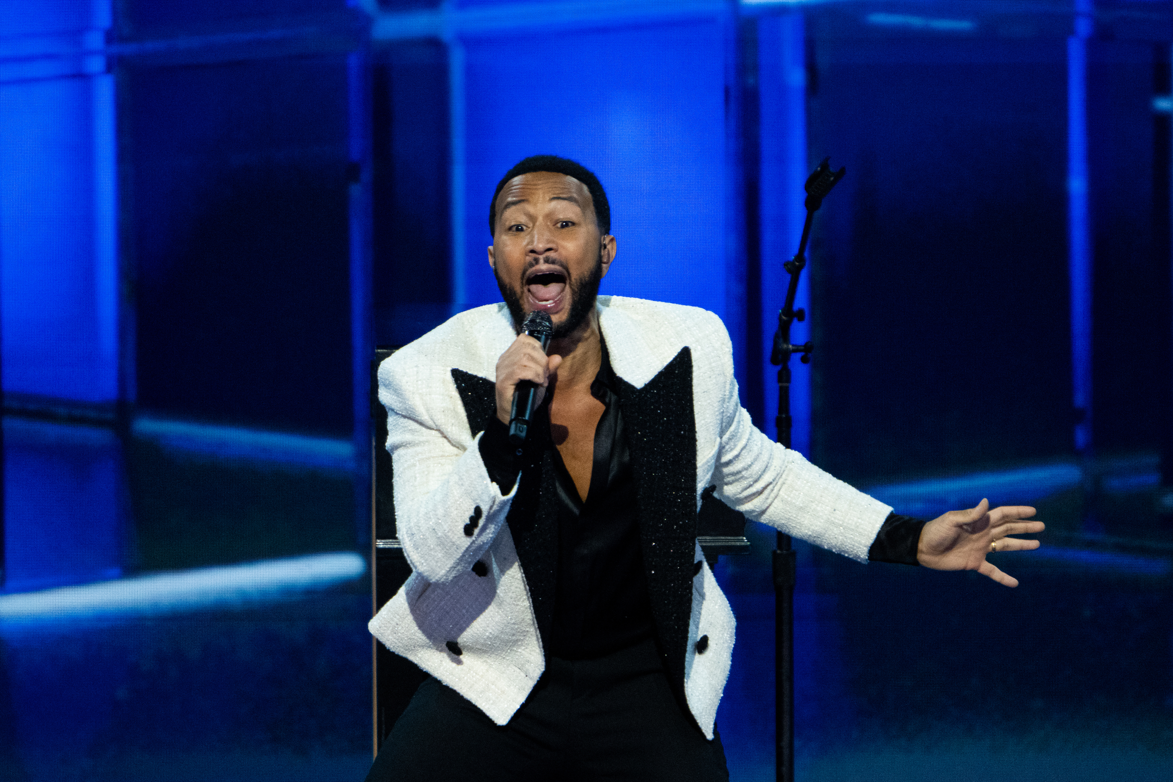 John Legend singing passionately on stage, wearing a white blazer with black details, a black shirt, and black pants