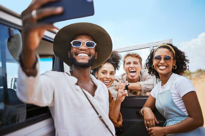 A group of friends, including two men and two women, happily take a selfie next to a car. They are enjoying a sunny day on a travel adventure