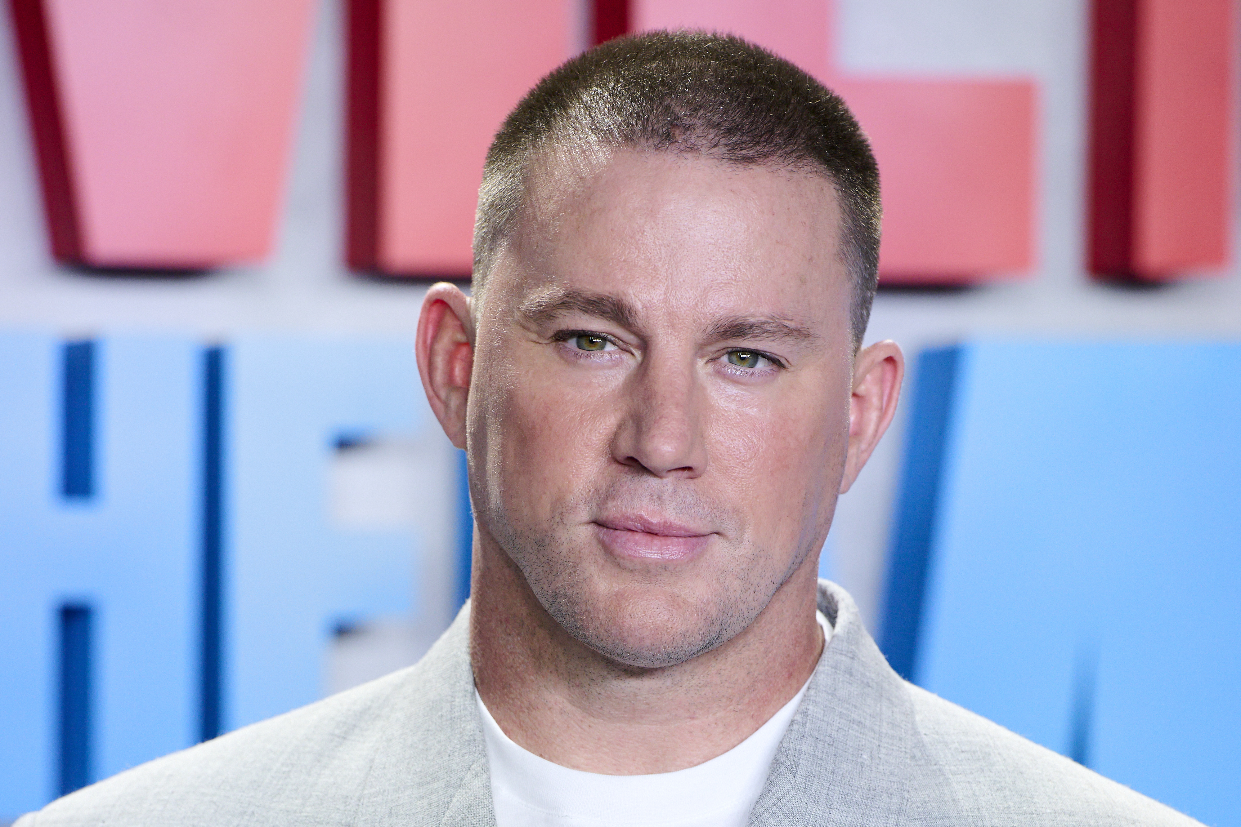 Channing Tatum is pictured in front of a backdrop with large text. He is wearing a light grey blazer over a white shirt