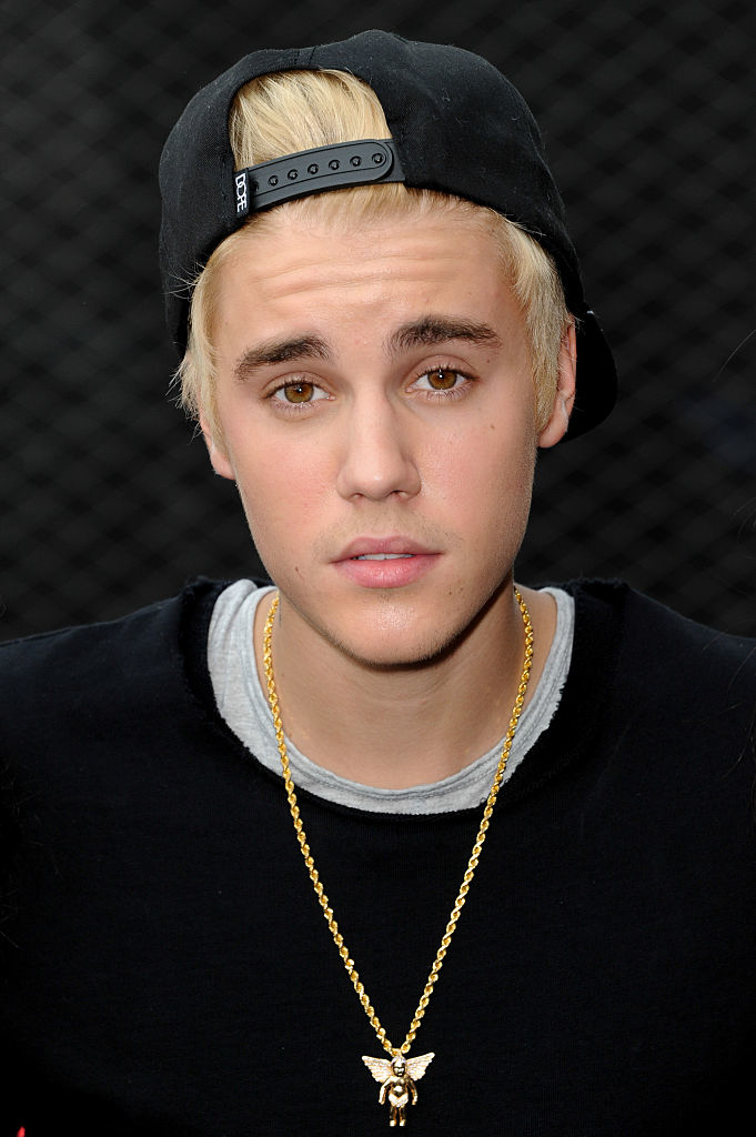 Justin Bieber wearing a black cap backwards, a black shirt, and a gold chain necklace, looking directly at the camera