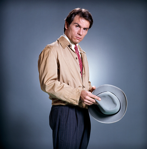 Sam Neill stands in a formal pose, wearing a beige jacket and striped trousers, holding a gray hat in his right hand against a plain background