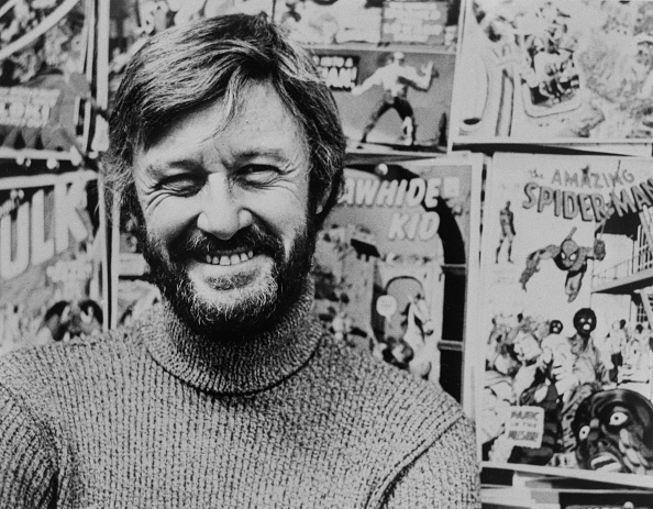 A man is smiling in front of a wall covered with comic book posters, including "The Amazing Spider-Man," "Rawhide Kid," and "Hulk"