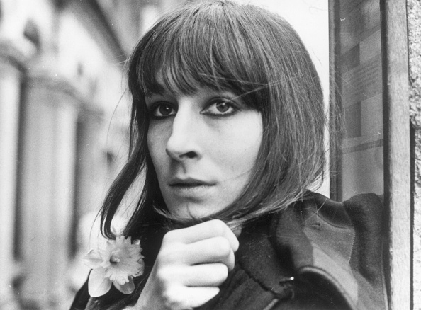 celeb poses close to a wall, holding a flower-shaped accessory near her coat, she gazes directly at the camera, showcasing her bangs and straight hair