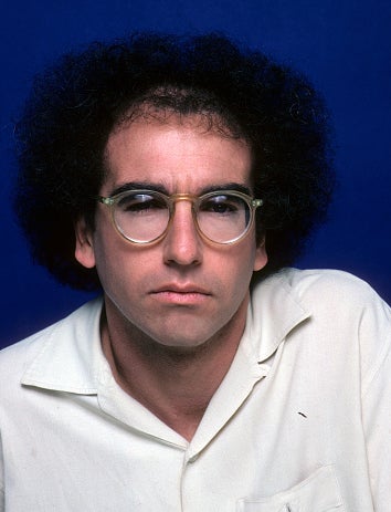 A person with curly hair and glasses wears a light-colored shirt and has a serious expression, posing against a plain background