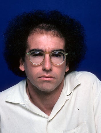A person with curly hair and glasses wears a light-colored shirt and has a serious expression, posing against a plain background