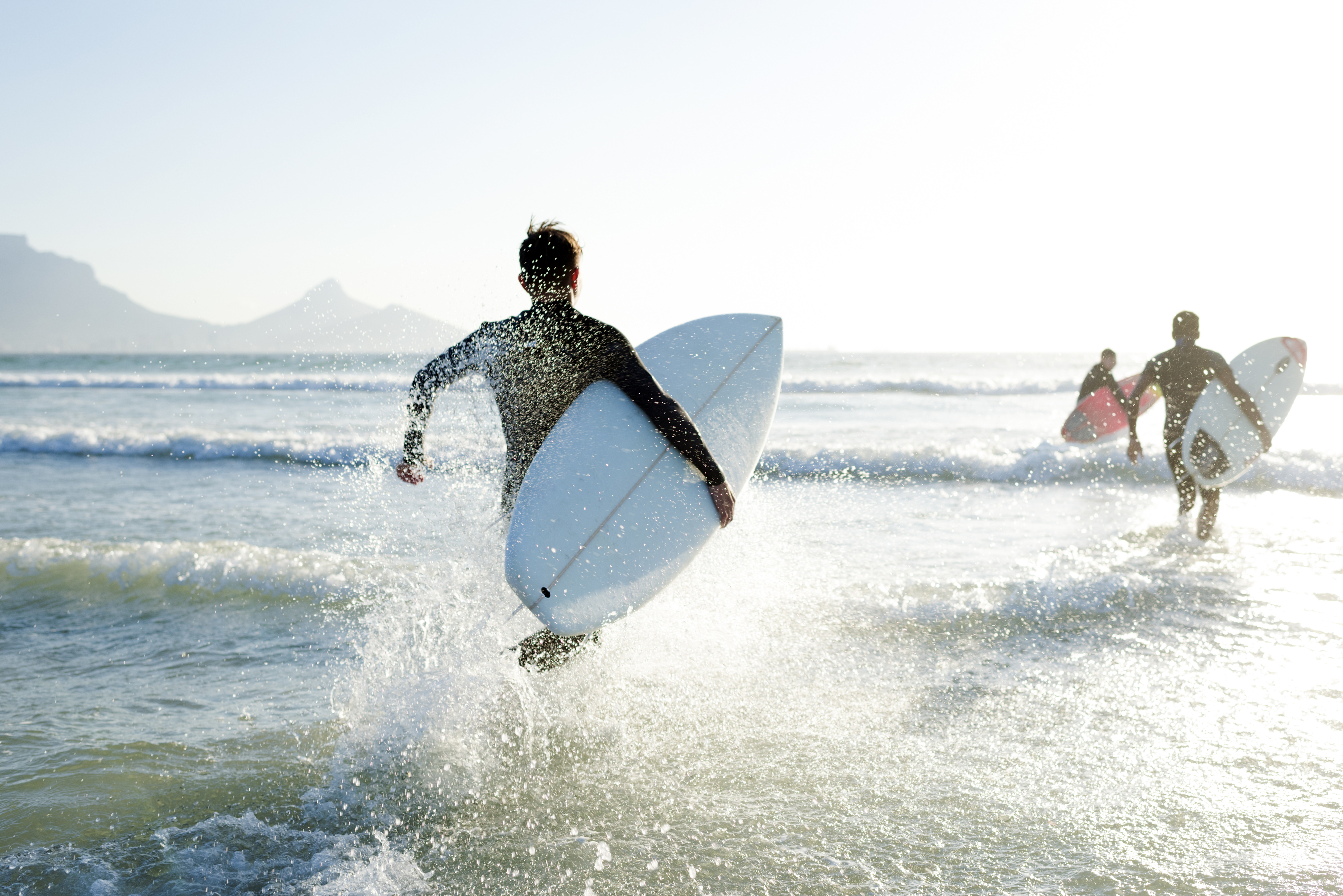 Three surfers, carrying their surfboards, run into the ocean waves