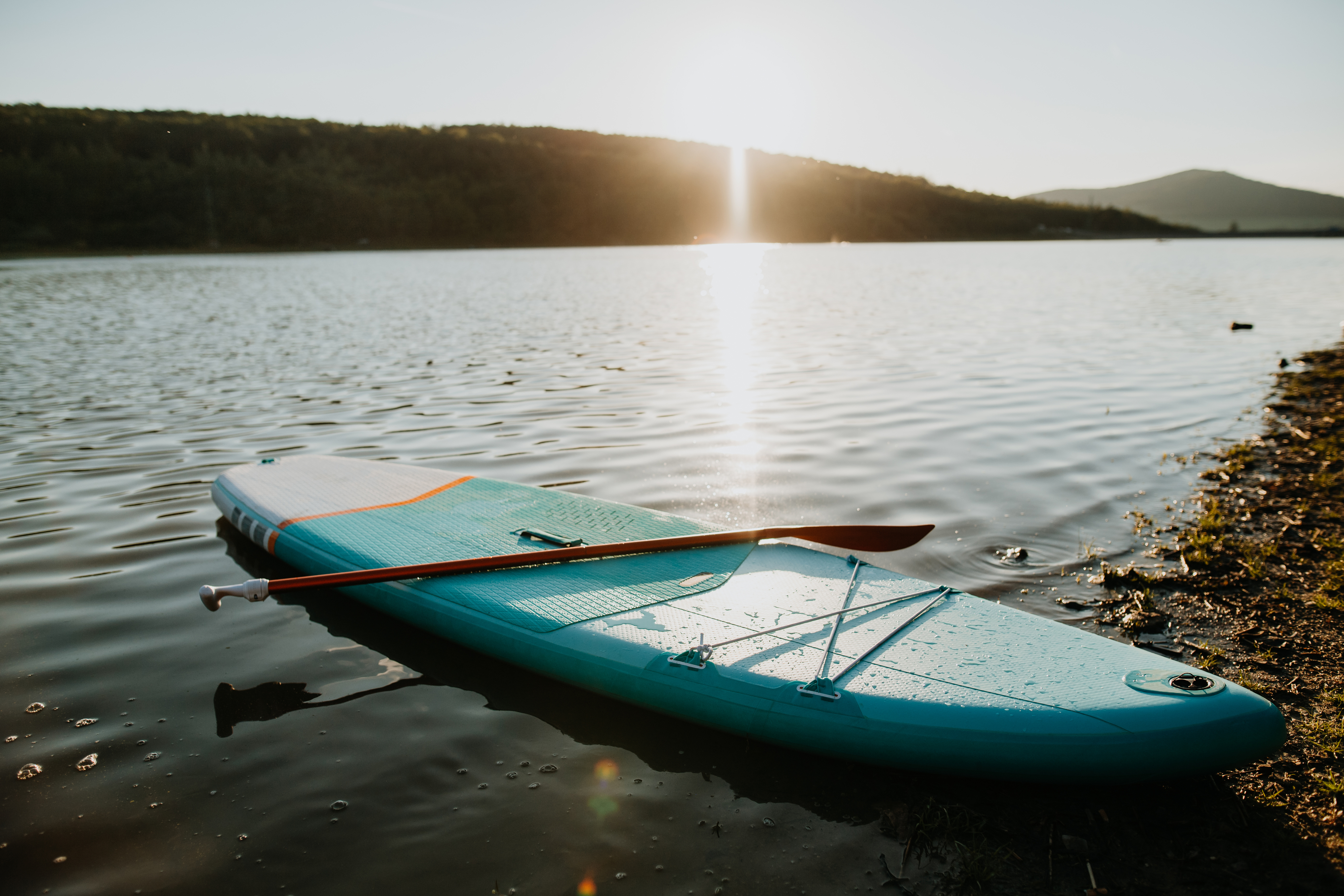 A paddleboard with an oar rests on the shore of a calm lake during sunset, surrounded by hills