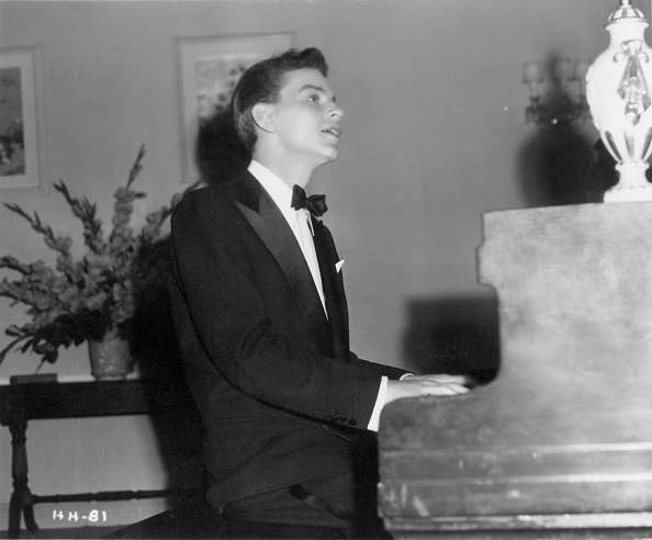 A man in a tuxedo is playing a piano with a vase on top, looking upwards possibly singing, the room has a potted plant in the background