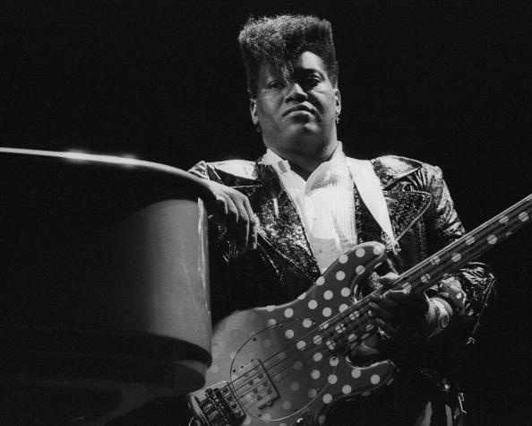 Man stands with an electric bass guitar, wearing a sparkling jacket and a white shirt