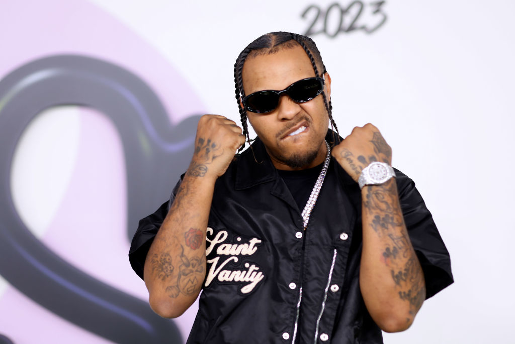 A man wearing black sunglasses and a black shirt with "Saint Vanity" printed on it poses with clenched fists. He has tattoos on both his arms